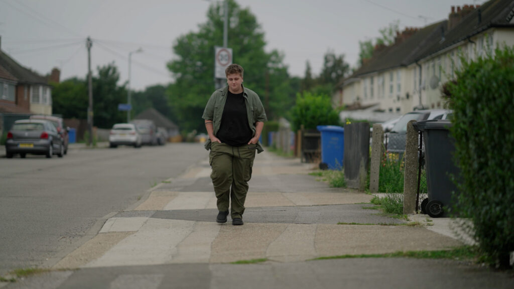 Jed walks towards the camera along the pavement of a residential street with his hands in his pockets.