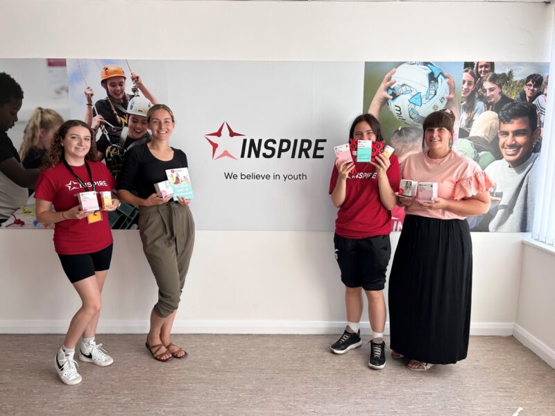 Inspire staff stand in front of a sign with an Inspire logo holding Hey Girls Period Products