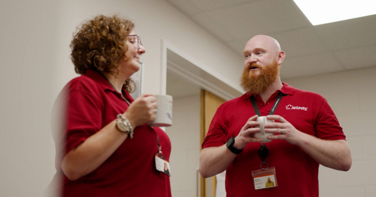 Two Youth Employment Service Coaches standing together in a hallway having a conversation while holding coffee mugs.