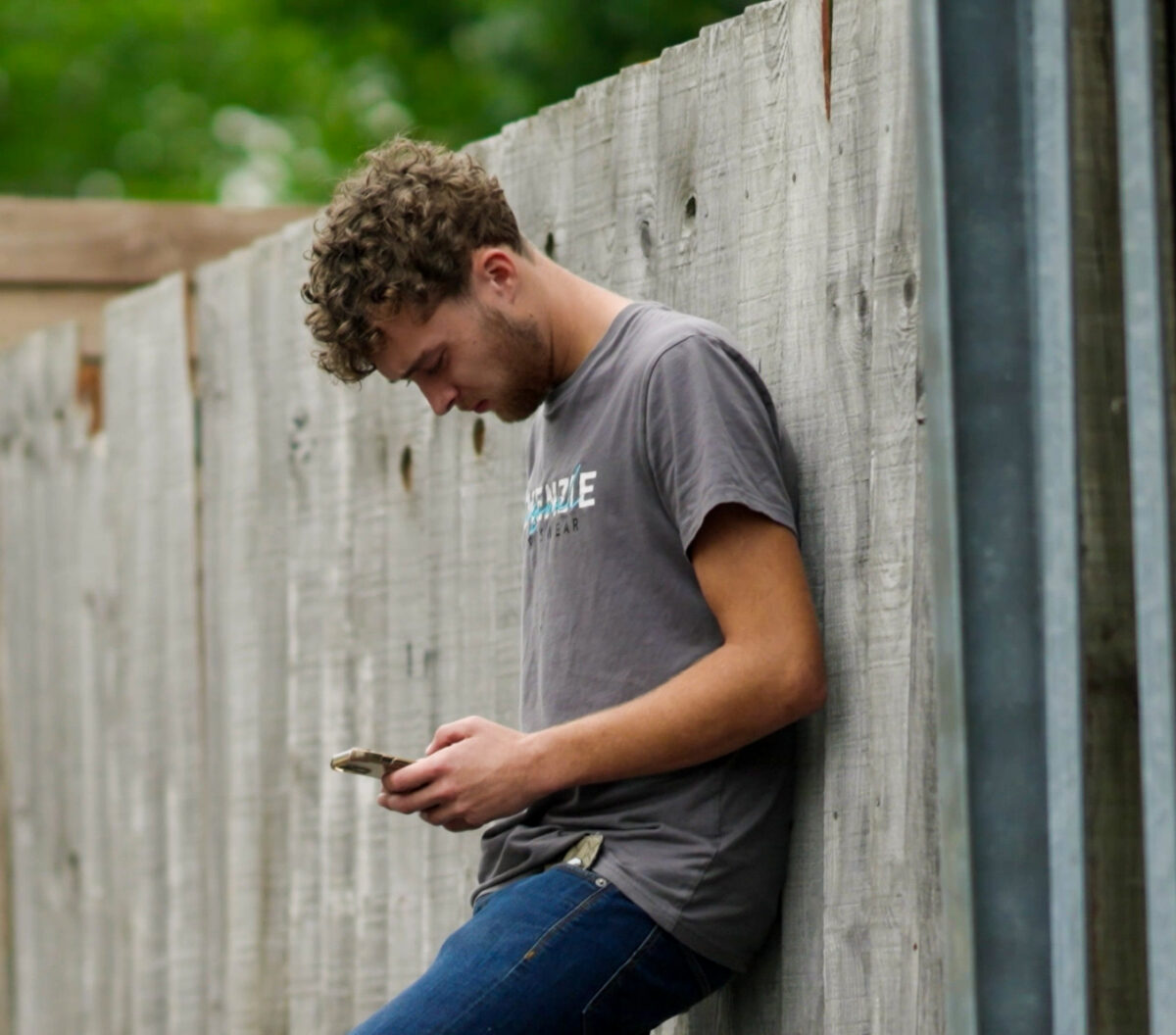 A young person in a grey t-shirt leans against a fence, checking their phone.